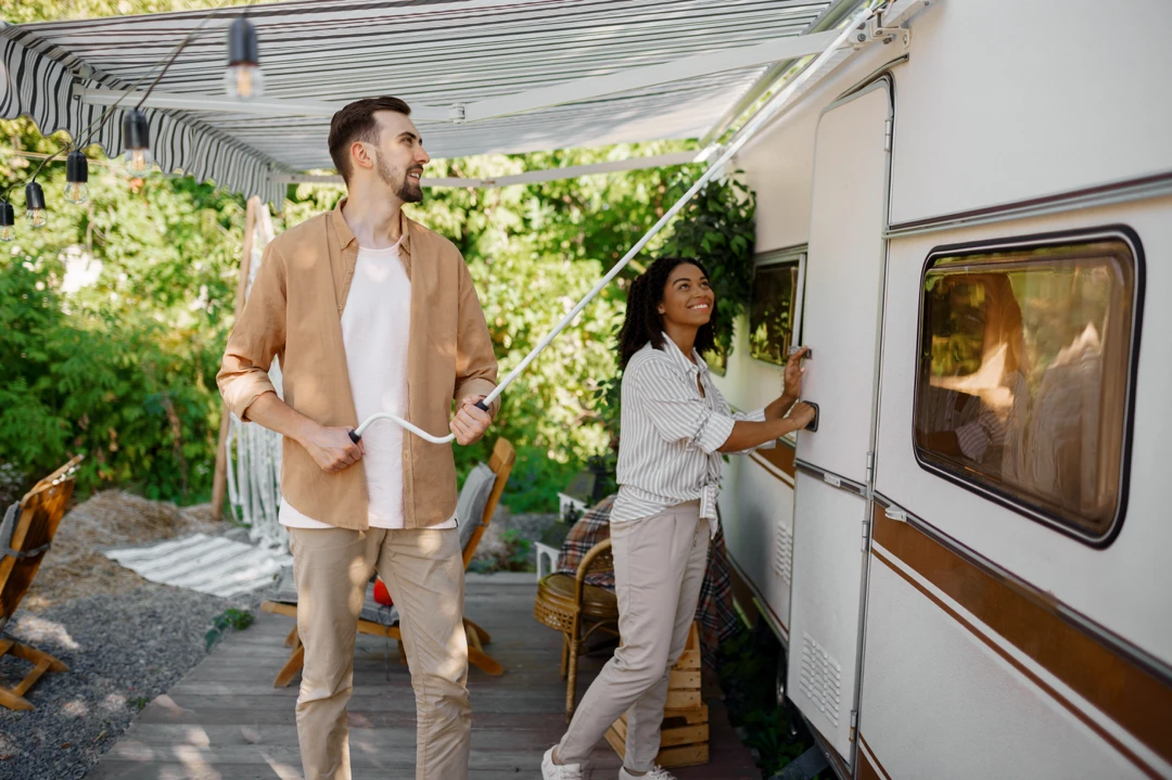 Couple installant un nouvel auvent sur une roulotte dans un terrain de camping en été, sous un ciel ensoleillé, entouré de verdure luxuriante.