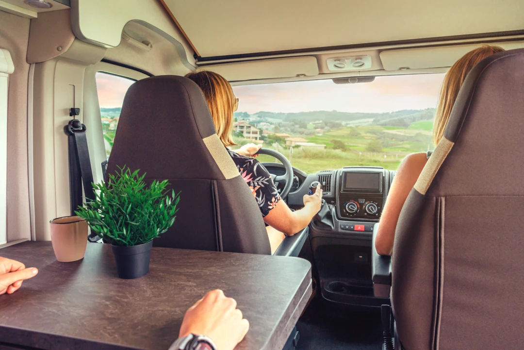 Femme au volant d’un VR roulant près du Lac-Saint-Jean, accompagnée d’une passagère et d’un passager, ambiance chaleureuse de road trip estival avec vue sur la campagne québécoise.