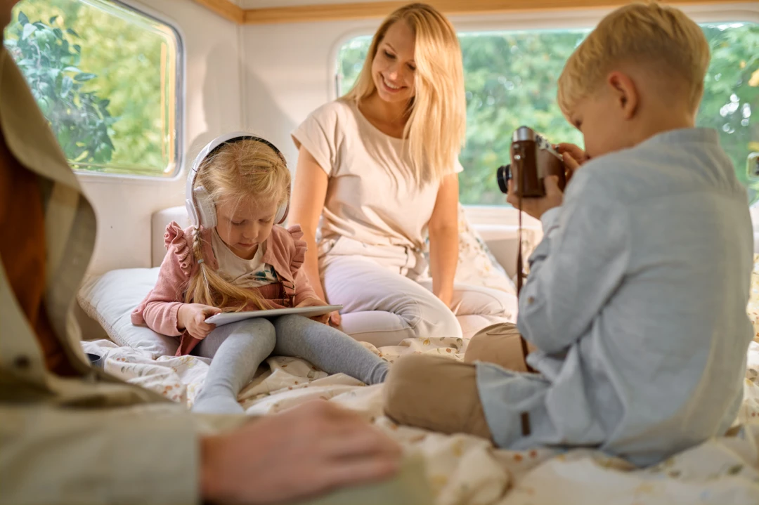 Scène familiale à l’intérieur d’une roulotte spacieuse : fillette avec écouteurs sur la tête concentrée sur une tablette, pendant que son frère la prend en photo sous le regard bienveillant de leur mère souriante.